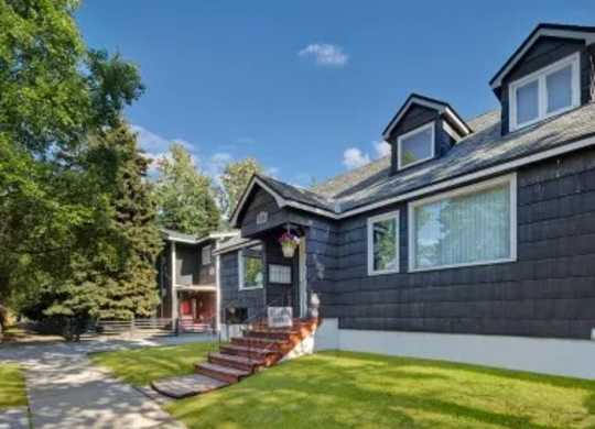 The leasing office at The Alaskan with dark siding, white trim, and welcoming front steps leading to the entrance. A large front window looks out onto a well-kept lawn with mature trees and a sidewalk, creating a friendly neighborhood feel on a bright day.
