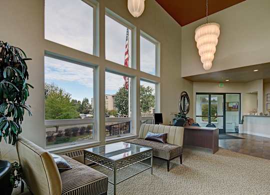 A bright lobby at The Alpine with tall windows letting in natural light and views of the community. Stylish seating surrounds a glass-top coffee table, and a desk sits near the entrance. Modern lighting, warm carpeting, wall art, and indoor plants create an inviting welcome area.