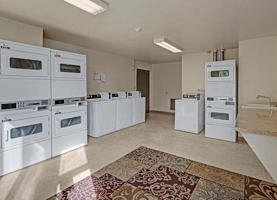 Spacious community laundry room at The Alpine featuring multiple front-loading washers and dryers, including stacked units, a folding counter with sink, and vending-style detergent machine. The bright space has neutral walls, overhead fluorescent lighting, and patterned area rug near the entrance.