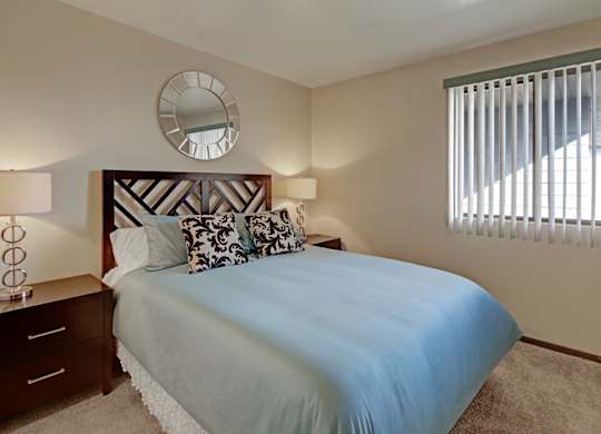 A cozy bedroom at The Alpine featuring a neatly made bed with blue bedding and patterned accent pillows. Matching bedside tables with lamps sit on each side of the headboard, and a window with vertical blinds brings in natural light. A round wall mirror adds a stylish touch above the bed.