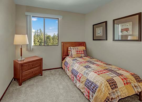 A bright bedroom at The Alpine featuring a twin bed with colorful patchwork bedding and a wooden headboard. A nightstand with a lamp sits near a large window that brings in natural light and views of the trees. Framed playful artwork adds a cozy, inviting touch.