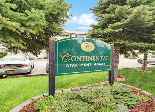 A welcoming community sign here at The Continental displaying Continental Apartment Homes branding, set within a landscaped green lawn with mature evergreen trees, surrounding shrubs, and nearby parking under a bright sky.