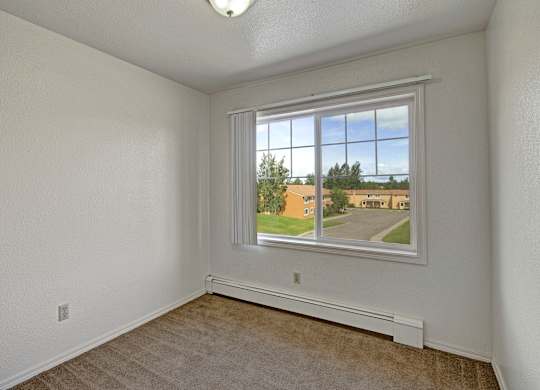 A bright and cozy bedroom here at The Glen featuring neutral walls, plush carpet flooring, and a large window with vertical blinds that fills the space with natural light while offering views of the surrounding community, creating a comfortable and versatile room ideal for a bedroom, home office, or personal retreat.