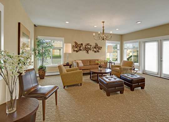 A welcoming community lounge here at The Glen featuring soft neutral walls, plush patterned carpet, and a coordinated seating area with sofas, armchairs, and ottomans arranged around a central coffee table, complemented by large windows, glass doors, warm lighting, and tasteful décor that creates a comfortable space for relaxing or gathering.