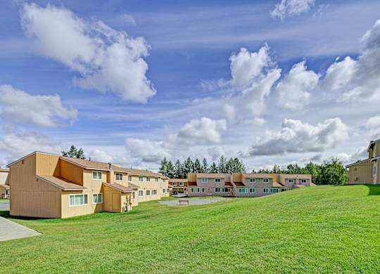 A wide outdoor view here at The Glen featuring tan two-story apartment buildings arranged around an open grassy courtyard, with a gently sloping lawn in the foreground, a small playground area in the center, mature trees lining the property, and a bright blue sky filled with soft white clouds overhead.
