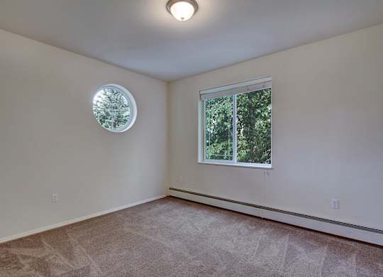 An empty and spacious bedroom here at The Highlands Luxury, with a small rounded window and a rectangular window on the right-most wall. With a soft singular light above on the ceiling..