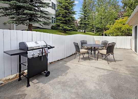 An outdoor amenity space here at The Highlands Luxury, including a barbecue grill, with a rounded outdoor table and chairs, sournded by white fences and concrete flooring.