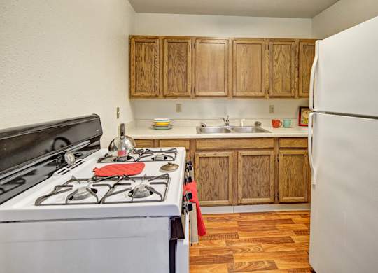 A cozy kitchen at The Village at Calais with wood cabinets, a white stove and refrigerator, a double sink, and wood-look flooring, accented by small colorful dishes and towels on the counters.
