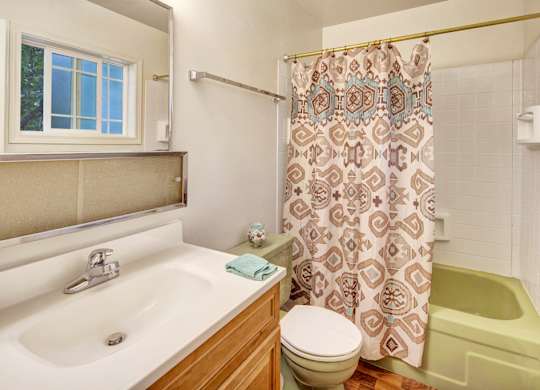 A compact bathroom at The Village at Calais with a wood vanity and white countertop, large mirror, toilet, and a tub/shower combo featuring a patterned shower curtain, white tile surround, and a small window bringing in natural light.