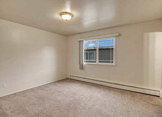 A spacious, empty bedroom here at The Village at Calais with soft beige carpet, clean white walls, and a wide window with vertical blinds that lets in natural light.