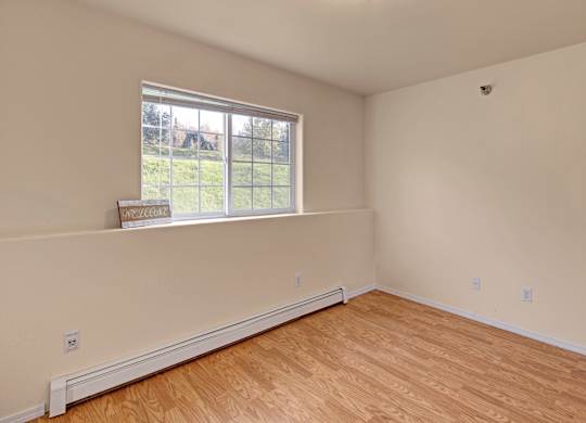 A bright, empty bedroom here at Timber Ridge with light wood-style flooring, soft neutral walls, and a large window bringing in natural light with a peaceful green view outside.
