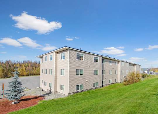 A sunny exterior view here at Timber Ridge showcasing a multi-story apartment building with neutral siding, large windows, and a spacious grassy lawn, surrounded by scenic trees under a bright blue sky.