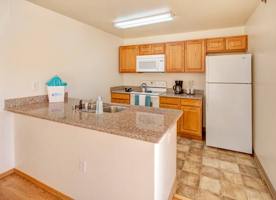 A bright kitchen here at Timber Ridge with warm wood cabinets, speckled granite-style countertops, and a stainless steel sink built into a large breakfast bar, featuring white appliances and a clean, open layout with tiled flooring.