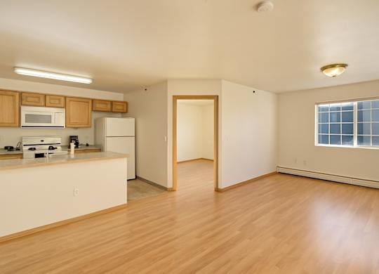 A spacious, open living area here at Timber Ridge with light wood flooring, a large window for natural light, and an open kitchen featuring warm wood cabinets, light countertops, and white appliances for a bright, inviting layout.