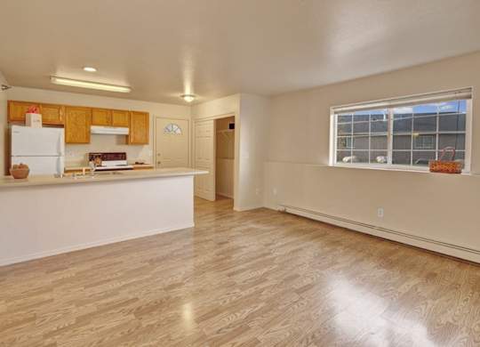 A spacious, empty living area here at Timber Ridge with wood-style flooring, soft neutral walls, and an open kitchen with warm wood cabinets, a wide breakfast bar, and white appliances, plus a large window bringing in natural light.