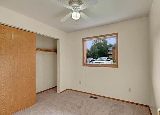 A bright, empty bedroom here at Wildwood Estates with soft carpet, a ceiling fan with overhead light, and a spacious closet with sliding doors, plus a window overlooking the community.
