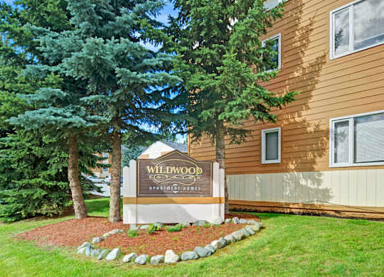 A welcoming community sign here at Wildwood Estates surrounded by mature evergreen trees, manicured grass, and neat landscaping in front of the apartment buildings.