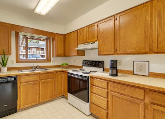 A bright kitchen here at Wildwood Estates with warm wood cabinets, light countertops, a stainless steel sink, and a dishwasher, featuring a spacious layout with a stove and a window for natural light.