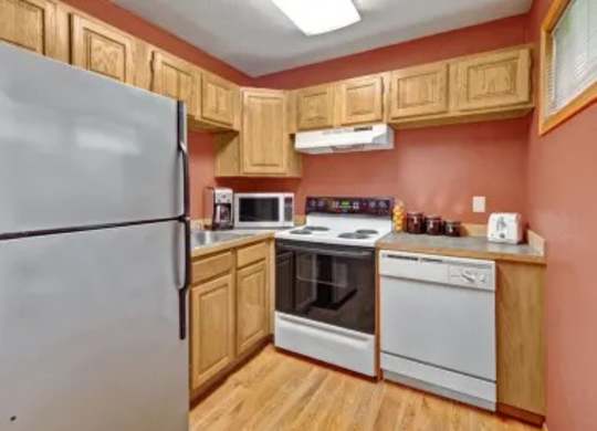 A cozy kitchen here at Wildwood Estates with warm wood cabinets, a full-size refrigerator, a stove, and a dishwasher, accented by rich terracotta walls and bright overhead lighting.