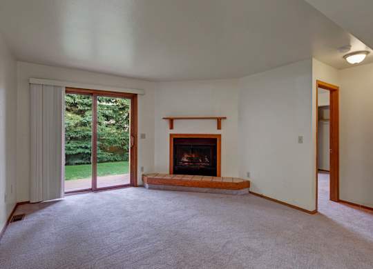 A spacious, empty living room here at Wildwood Estates with soft carpet, a cozy fireplace with a tiled hearth, and a sliding glass door that opens to a patio with peaceful green views.