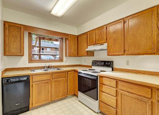 A bright kitchen here at Wildwood Estates with warm wood cabinets, light countertops, a stainless steel sink, and a dishwasher, featuring a stove and a large window that brings in natural light.