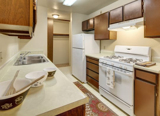 Narrow kitchen here at Taiga Apartments with brown cabinets, white appliances including a refrigerator and gas stove, three bowls on the counter, and a patterned rug on the floor.