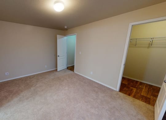 Beige Bedroom here at Four Seasons Apartments with carpeted floor, lit by a ceiling light. Open doors reveal a hallway with wooden flooring and a closet with wire shelving.