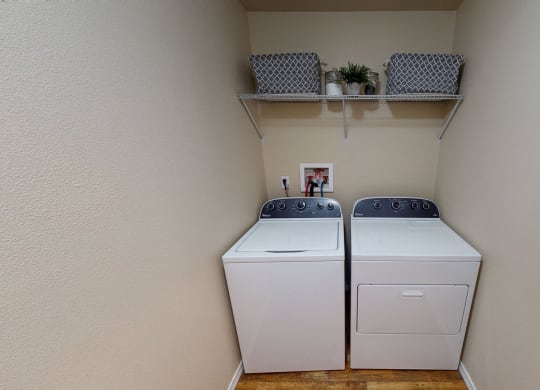 Laundry room here at Four Seasons Apartments with a washer and dryer side by side. Above them is a shelf with two patterned baskets and plants. The setting feels tidy and organized.