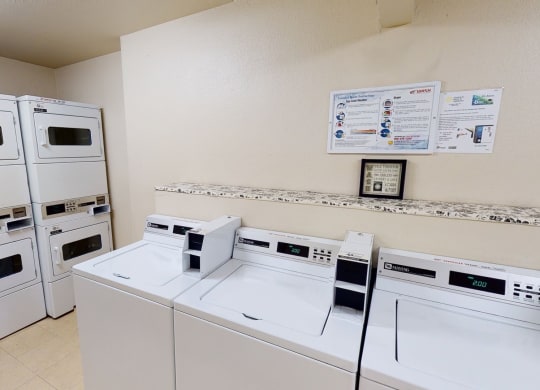 A clean laundry room here at Hillside Chalet Apartments with stacked dryers on the left and top-loader washers on the right. Instructions are mounted on a beige wall, creating an orderly atmosphere.