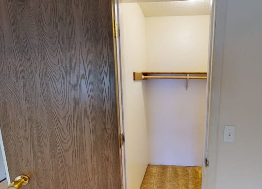 Open closet here at Rancho Tudor Apartments with wooden door ajar, revealing a white interior, a wooden hanging rod, and beige carpet. The space is minimal and tidy.