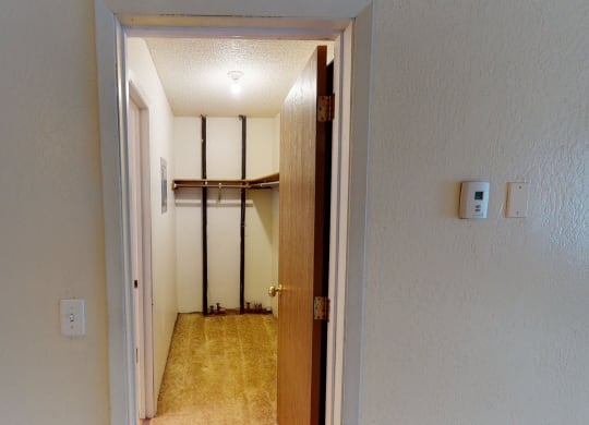 A small, empty closet here at Rancho Tudor Apartments with beige walls and carpet, an open wooden door, a single light bulb, metal rods, wooden shelves, and a wall thermostat.