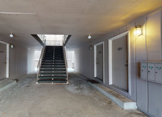 Apartment hallway here at Rancho Tudor Apartments with beige walls and closed brown doors numbered 403, 410, 409, 404. Central staircase leads to upper floor. Lit by small ceiling lights. Calm atmosphere.