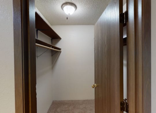 A small, empty walk-in closet here at Terrace on the Lake Apartments with beige carpet, white walls, and wooden shelves on the left. An open wooden door and a ceiling light create a cozy ambiance.
