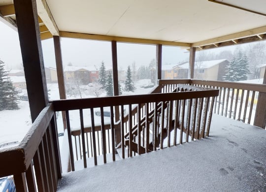 Snow-covered porch here at The Club at Eagle Pointe Apartments with wooden railings overlooks a snowy neighborhood. Fir trees and houses are visible under a gray, overcast sky. Quiet, wintry scene.