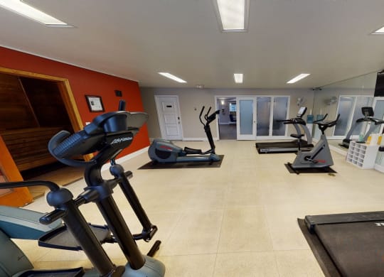 Bright gym room here at The Club at Eagle Pointe Apartments featuring treadmills, ellipticals, and stationary bikes on tiled floor. Mirrors and a flatscreen TV create a modern, spacious feel.