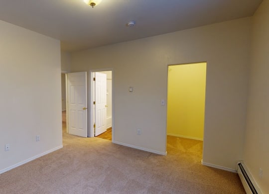Empty bedroom here at The Highlands Luxury Apartments with beige walls and carpet, featuring three open white doors leading to a bathroom and hallway. Soft lighting creates a calm atmosphere.