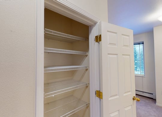 Open white closet door revealing empty wire shelves inside a beige wall alcove. Bright room here at The Highlands Luxury Apartments with carpet and window in the background.