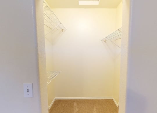 Walk-in closet here at The Highlands Luxury Apartments with beige walls, carpeted floor, and wire shelving on three sides. A single ceiling light provides a bright, clean ambiance.