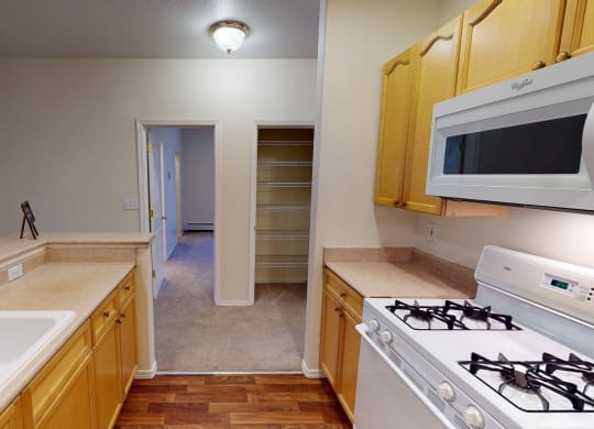 Kitchen interior here at The Highlands Luxury Apartments with light wood cabinets, a gas stove, white microwave, and double sink. Hallway with open pantry on the right leads to another room.