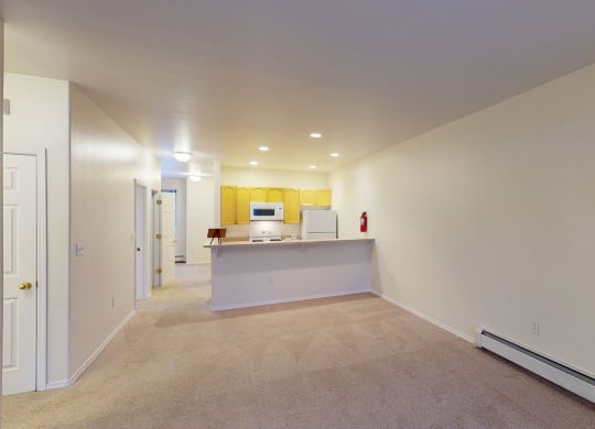 Spacious, empty living room here at The Highlands Luxury Apartments with beige carpet and walls, leading to a kitchen with yellow cabinets, white appliances, and warm ceiling lights.
