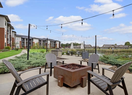 Outdoor patio here at 49 West Apartments with Adirondack chairs around a square fire pit. String lights hang above a pathway, leading to modern apartments and lush greenery. Relaxing ambiance.