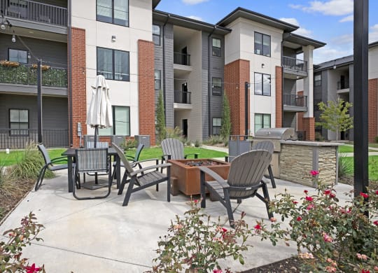 Modern 49 West Apartments courtyard with patio furniture, grill, and fire pit. Surrounding buildings are multi-story with brick and beige exteriors. Lush greenery adds a welcoming feel.