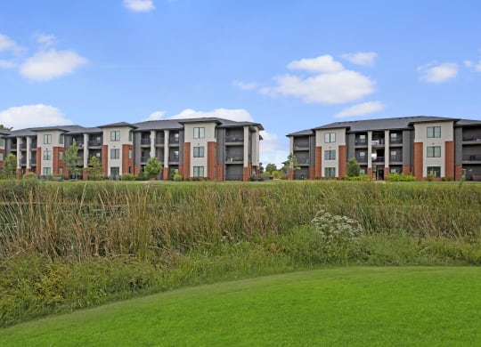Modern three-story 49 West Apartment buildings with a mix of beige and brick exteriors sit behind a lush wetland, under a clear blue sky.