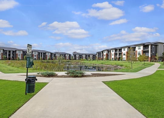 A serene view of modern 49 West Apartment buildings surrounding a central pond under a blue sky with clouds. Green lawns and pathways create a welcoming atmosphere.