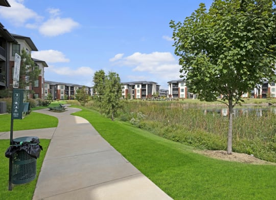 Paved path curves through a green park, lined with trees near 49 West Apartment buildings. Bright blue sky with clouds creates a serene and inviting atmosphere.