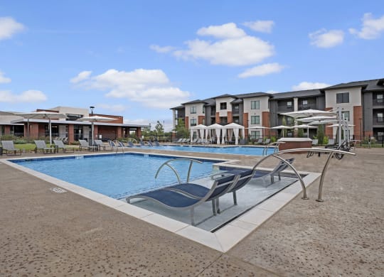 Spacious outdoor pool area here at 49 West Apartments with modern lounge chairs and umbrellas, surrounded by a residential complex. The scene is bright and inviting under a clear blue sky.