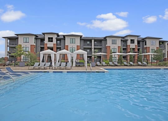 Luxurious poolside view of 49 West's modern apartment complex, featuring cabanas and lounge chairs under a blue sky with fluffy clouds. Relaxing and inviting atmosphere.