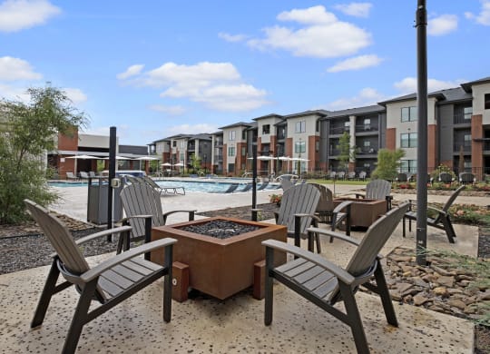 Modern 49 West Apartment courtyard featuring a fire pit surrounded by Adirondack chairs. A pool and lounge chairs are visible under a blue sky. Relaxed ambiance.