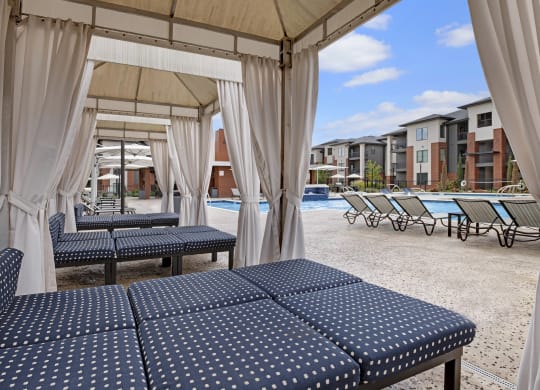 Poolside cabana here at 49 West Apartments with polka-dotted lounge chairs, surrounded by white curtains, overlooking a pool and modern apartment buildings under a blue sky.