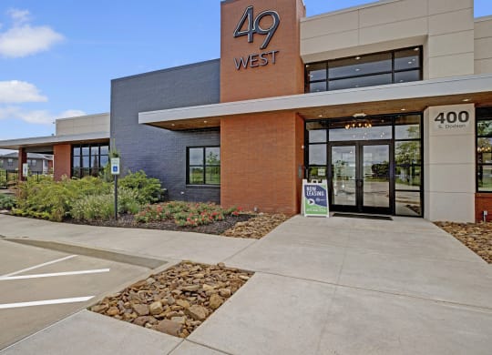 Modern building exterior with "49 West" sign, large glass entrance, and red brick facade. "Now Leasing" sign outside. Clear blue sky and greenery.